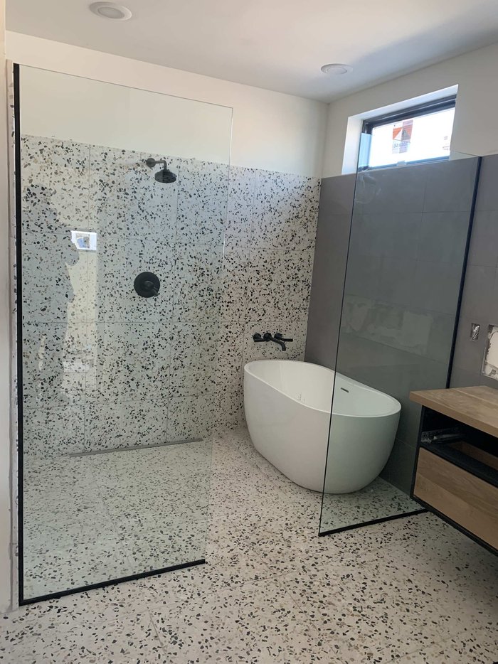 Frameless walk-in shower panels with matte black hardware and terrazzo tile beside a soaking tub